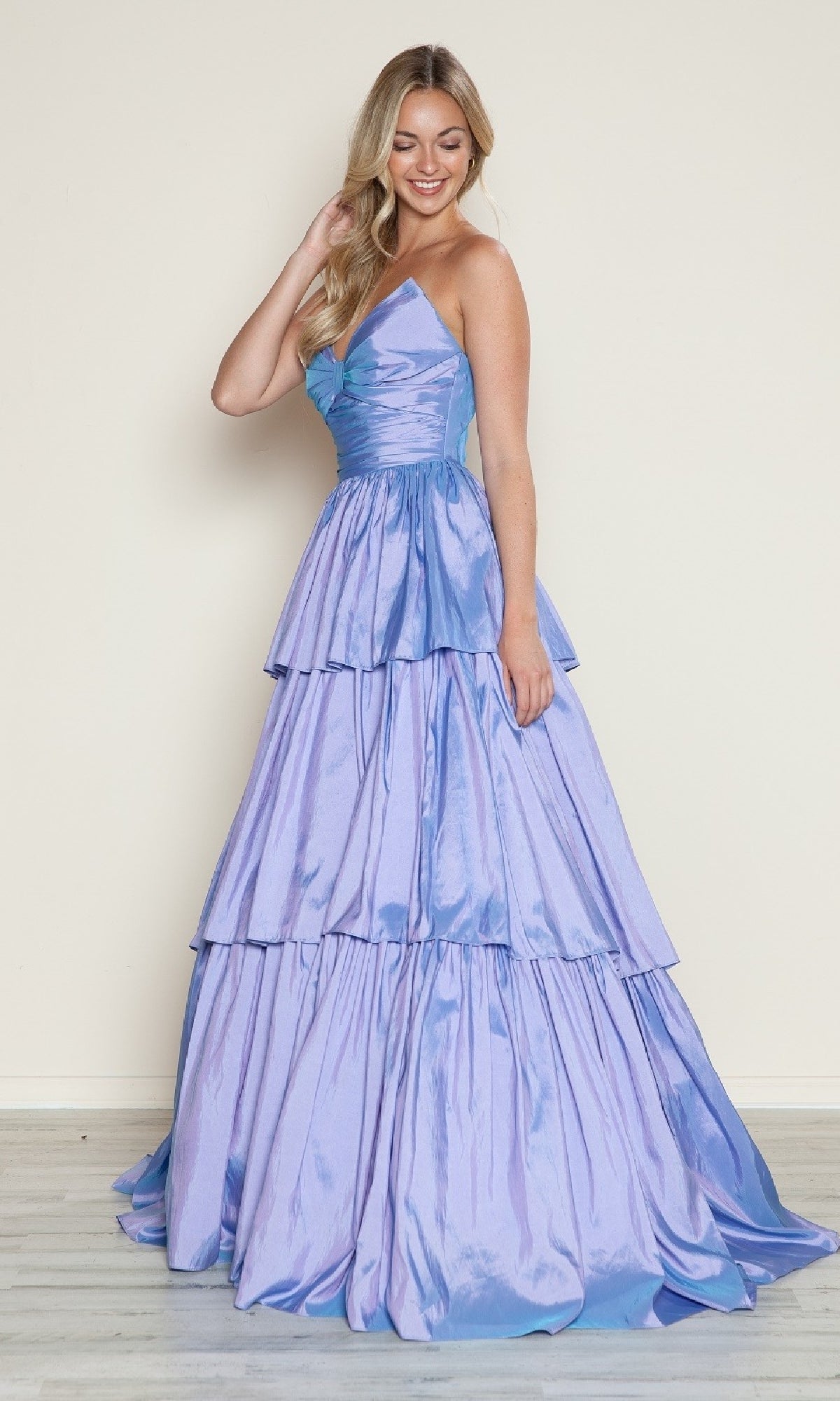 A woman smiles and touches her hair while wearing the Long Prom Dress: Poly USA 9694, a shiny, light purple strapless gown with tiered a-line layers, standing against a plain light background.